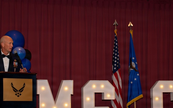 A senior U.S. Air Force enlisted member, identifiable by Chief Master Sergeant rank insignia on his mess dress uniform, delivers a speech at a formal military banquet. The event appears to be an MSGT (Master Sergeant) promotion ceremony or recognition dinner, indicated by illuminated marquee letters spelling 'MSGT' on stage. The setting includes an audience seated at formal dining tables, blue and black balloons, and both the U.S. flag and Air Force flag displayed on stage.