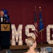 A senior U.S. Air Force enlisted member, identifiable by Chief Master Sergeant rank insignia on his mess dress uniform, delivers a speech at a formal military banquet. The event appears to be an MSGT (Master Sergeant) promotion ceremony or recognition dinner, indicated by illuminated marquee letters spelling 'MSGT' on stage. The setting includes an audience seated at formal dining tables, blue and black balloons, and both the U.S. flag and Air Force flag displayed on stage.