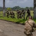 A group of military personnel in camouflage uniforms are conducting a tactical training exercise in a tropical outdoor environment. Several soldiers are moving in a crouched formation while carrying rifles, with one instructor or observer positioned in the foreground on a paved road. The setting appears to be a military training facility surrounded by palm trees and open grassland under overcast skies.