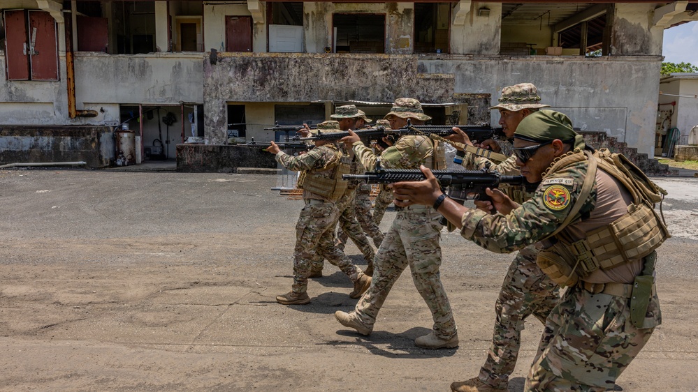 A group of military or law enforcement personnel in camouflage uniforms are conducting a tactical training exercise in an outdoor urban environment. They are moving in a line formation while aiming rifles, practicing a combat patrol or room-clearing drill. The setting appears to be a deteriorated building complex used as a training facility.