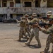 A group of military or law enforcement personnel in camouflage uniforms are conducting a tactical training exercise in an outdoor urban environment. They are moving in a line formation while aiming rifles, practicing a combat patrol or room-clearing drill. The setting appears to be a deteriorated building complex used as a training facility.