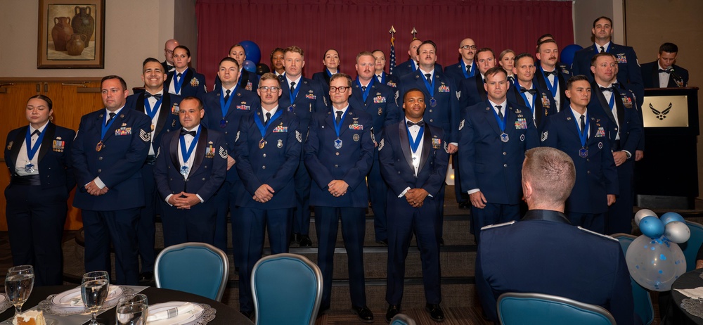A large group of U.S. Air Force personnel posed for a formal group photograph at what appears to be a military awards ceremony or banquet. The majority are wearing dark blue dress uniforms with blue medallion lanyards/medals around their necks, indicating they are honorees. The event is held in a banquet hall decorated with blue and silver balloons, with an American flag and Air Force symbol visible in the background.