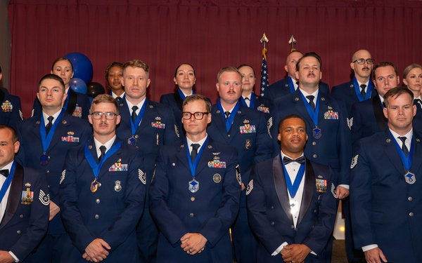 A large group of U.S. Air Force personnel posed for a formal group photograph at what appears to be a military awards ceremony or banquet. The majority are wearing dark blue dress uniforms with blue medallion lanyards/medals around their necks, indicating they are honorees. The event is held in a banquet hall decorated with blue and silver balloons, with an American flag and Air Force symbol visible in the background.
