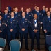 A large group of U.S. Air Force personnel posed for a formal group photograph at what appears to be a military awards ceremony or banquet. The majority are wearing dark blue dress uniforms with blue medallion lanyards/medals around their necks, indicating they are honorees. The event is held in a banquet hall decorated with blue and silver balloons, with an American flag and Air Force symbol visible in the background.