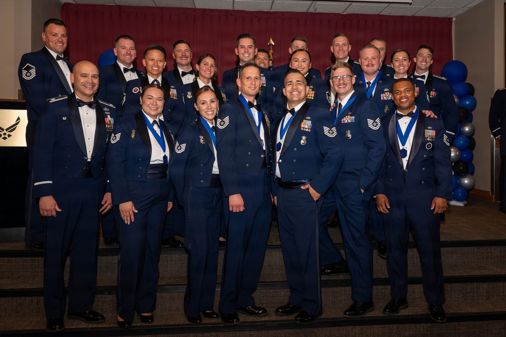 A large group of U.S. Air Force personnel posing for a formal group photograph at what appears to be an awards ceremony or military ball. The individuals are wearing dress blue uniforms and formal attire, with many displaying medals and ribbons on blue lanyards. The setting includes a decorated backdrop with maroon curtains, blue and silver balloons, and an Air Force emblem visible to the left.