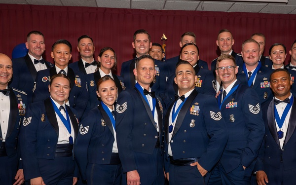A large group of U.S. Air Force personnel posing for a formal group photograph at what appears to be an awards ceremony or military ball. The individuals are wearing dress blue uniforms and formal attire, with many displaying medals and ribbons on blue lanyards. The setting includes a decorated backdrop with maroon curtains, blue and silver balloons, and an Air Force emblem visible to the left.