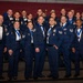 A large group of U.S. Air Force personnel posing for a formal group photograph at what appears to be an awards ceremony or military ball. The individuals are wearing dress blue uniforms and formal attire, with many displaying medals and ribbons on blue lanyards. The setting includes a decorated backdrop with maroon curtains, blue and silver balloons, and an Air Force emblem visible to the left.