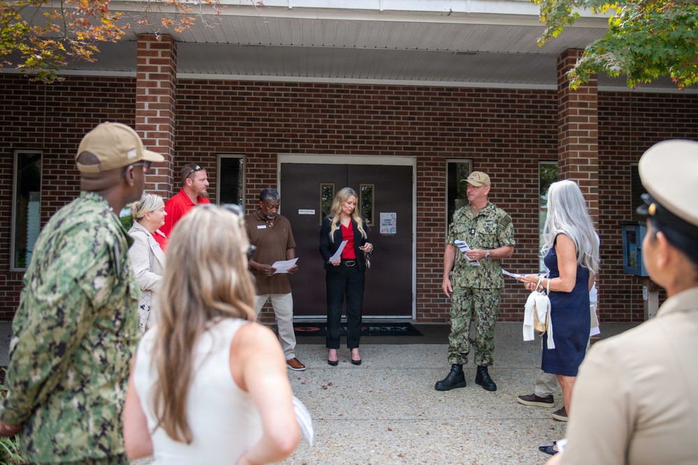 A small outdoor gathering is taking place in front of a brick building, involving both military personnel in camouflage uniforms and civilians in business casual attire. Several individuals appear to be reading from papers or documents, suggesting a formal ceremony, briefing, or presentation. The setting appears to be a military installation or government facility.
