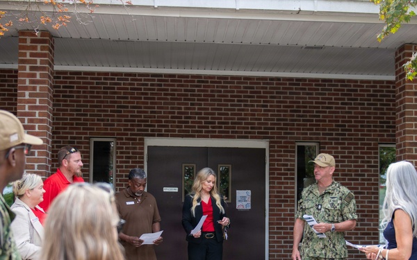 A small outdoor gathering is taking place in front of a brick building, involving both military personnel in camouflage uniforms and civilians in business casual attire. Several individuals appear to be reading from papers or documents, suggesting a formal ceremony, briefing, or presentation. The setting appears to be a military installation or government facility.