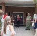 A small outdoor gathering is taking place in front of a brick building, involving both military personnel in camouflage uniforms and civilians in business casual attire. Several individuals appear to be reading from papers or documents, suggesting a formal ceremony, briefing, or presentation. The setting appears to be a military installation or government facility.
