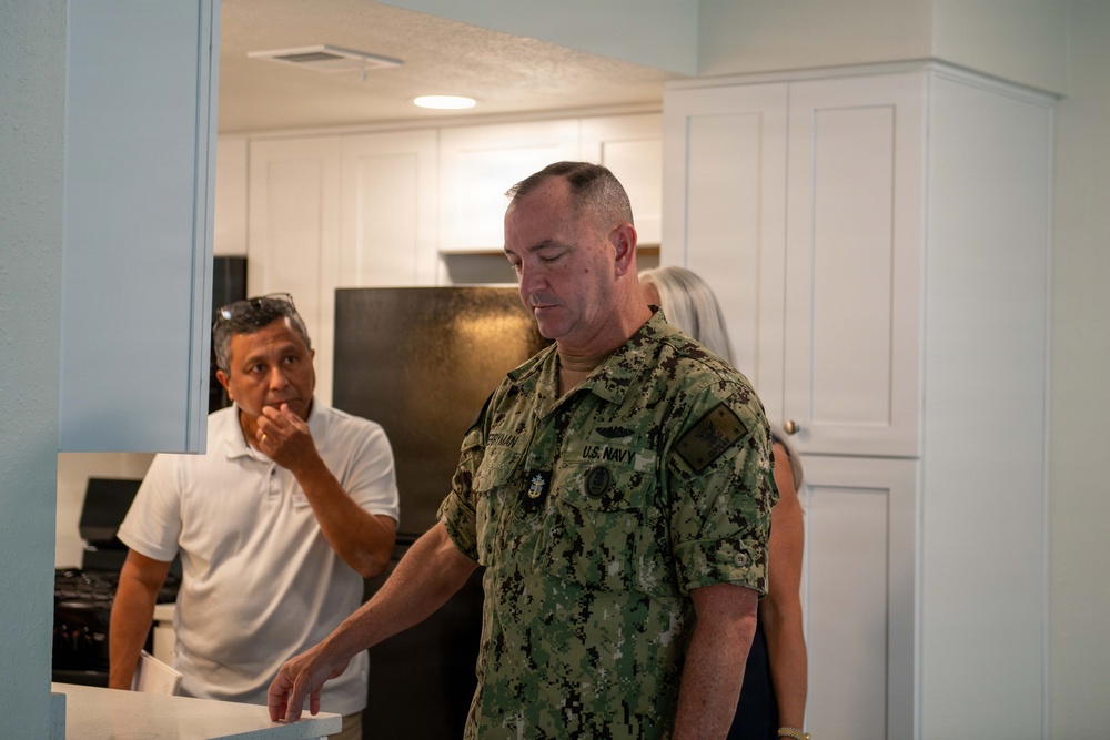 A U.S. Navy servicemember in NWU Type III digital camouflage uniform is standing in what appears to be a residential kitchen or housing unit, looking downward at a counter. He is accompanied by at least two civilians, one male in a white polo shirt and one partially visible female with gray hair. The setting suggests an inspection or tour of military housing facilities.