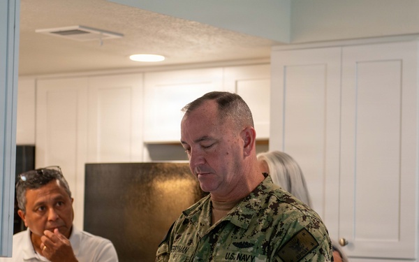 A U.S. Navy servicemember in NWU Type III digital camouflage uniform is standing in what appears to be a residential kitchen or housing unit, looking downward at a counter. He is accompanied by at least two civilians, one male in a white polo shirt and one partially visible female with gray hair. The setting suggests an inspection or tour of military housing facilities.