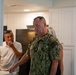 A U.S. Navy servicemember in NWU Type III digital camouflage uniform is standing in what appears to be a residential kitchen or housing unit, looking downward at a counter. He is accompanied by at least two civilians, one male in a white polo shirt and one partially visible female with gray hair. The setting suggests an inspection or tour of military housing facilities.