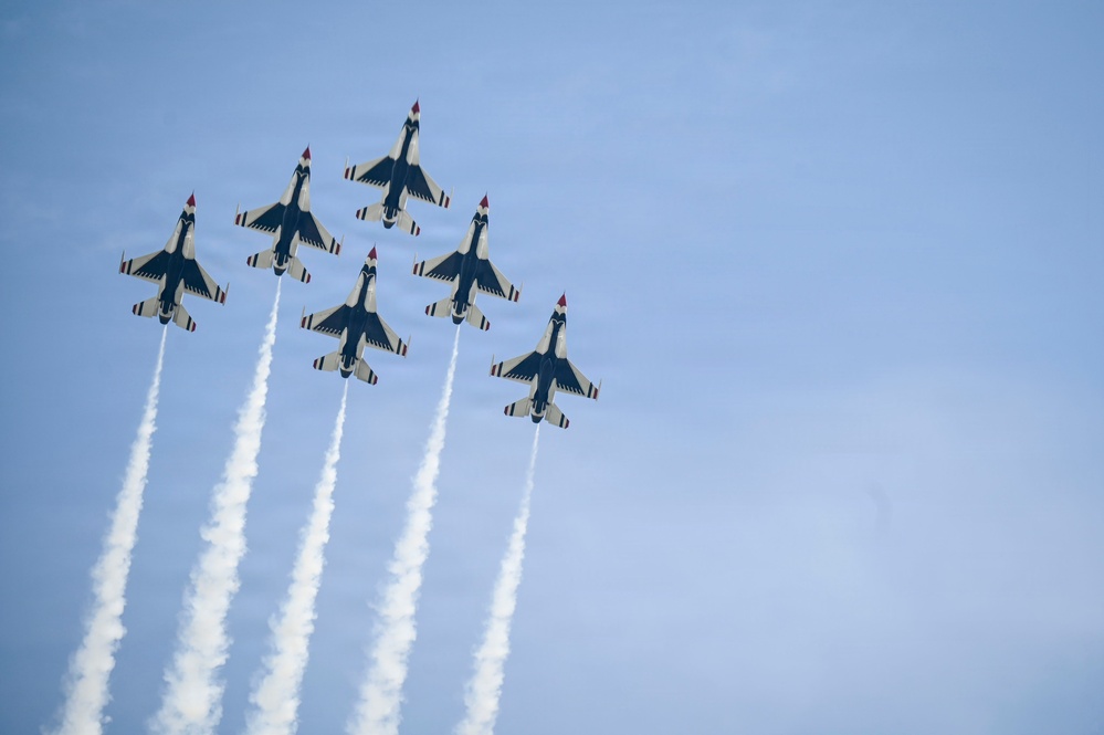 Six military jet aircraft are flying in a tight diamond formation against a clear blue sky, each trailing white smoke streams. The aircraft display a distinctive white, blue, and red color scheme consistent with the U.S. Air Force Thunderbirds aerial demonstration squadron. The formation and smoke trails suggest this is a precision aerobatic performance at an air show.