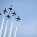 Six military jet aircraft are flying in a tight diamond formation against a clear blue sky, each trailing white smoke streams. The aircraft display a distinctive white, blue, and red color scheme consistent with the U.S. Air Force Thunderbirds aerial demonstration squadron. The formation and smoke trails suggest this is a precision aerobatic performance at an air show.