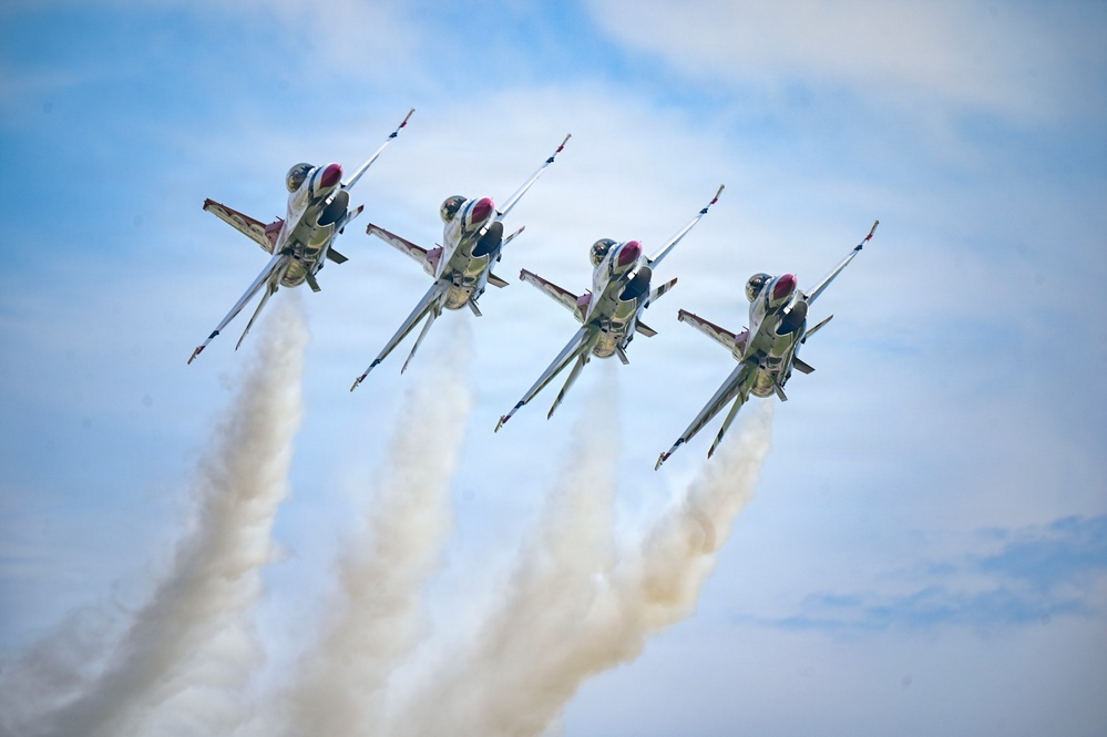 Four military jet aircraft are flying in a tight diamond formation against a partly cloudy blue sky. Each aircraft is producing a white smoke trail, creating four parallel contrails trailing behind the formation. The aircraft display red, white, and blue markings consistent with the U.S. Air Force Thunderbirds aerial demonstration team.