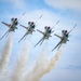 Four military jet aircraft are flying in a tight diamond formation against a partly cloudy blue sky. Each aircraft is producing a white smoke trail, creating four parallel contrails trailing behind the formation. The aircraft display red, white, and blue markings consistent with the U.S. Air Force Thunderbirds aerial demonstration team.