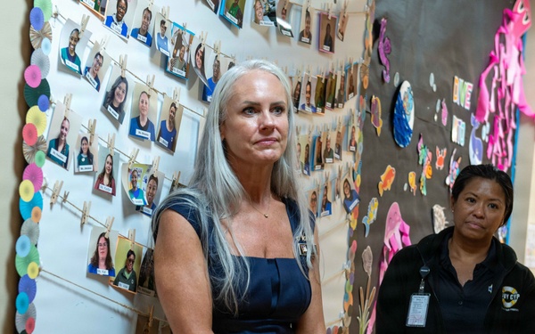 Two women stand in a decorated hallway or corridor, likely inside a school or community facility. The foreground woman has long gray hair and wears a navy dress while holding a notebook, and appears to be listening or observing. Behind her, a second woman in a black uniform jacket with a badge/ID lanyard stands attentively.
