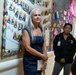 Two women stand in a decorated hallway or corridor, likely inside a school or community facility. The foreground woman has long gray hair and wears a navy dress while holding a notebook, and appears to be listening or observing. Behind her, a second woman in a black uniform jacket with a badge/ID lanyard stands attentively.