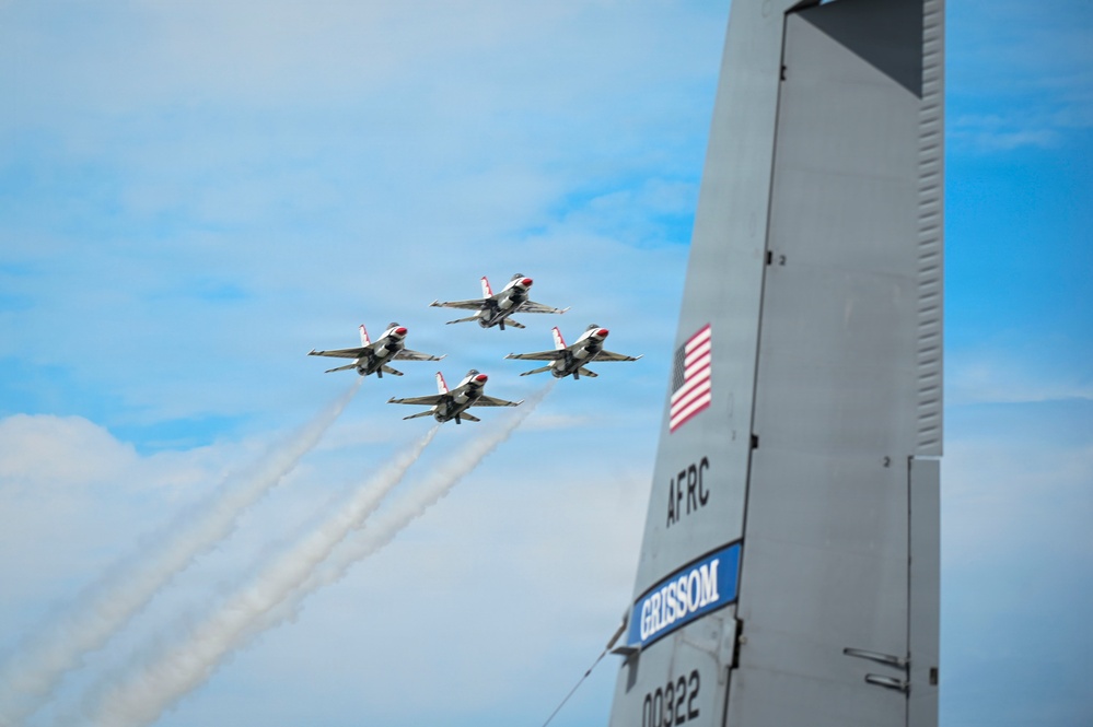 Four U.S. Air Force Thunderbirds aerobatic demonstration team jets fly in a tight diamond formation, leaving white smoke trails against a partly cloudy blue sky. In the foreground, the vertical tail fin of a large military aircraft is visible, marked with 'AFRC', 'GRISSOM', and the serial number '00322', along with a U.S. flag decal. This appears to be an air show at or associated with Grissom Air Reserve Base.