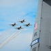 Four U.S. Air Force Thunderbirds aerobatic demonstration team jets fly in a tight diamond formation, leaving white smoke trails against a partly cloudy blue sky. In the foreground, the vertical tail fin of a large military aircraft is visible, marked with 'AFRC', 'GRISSOM', and the serial number '00322', along with a U.S. flag decal. This appears to be an air show at or associated with Grissom Air Reserve Base.