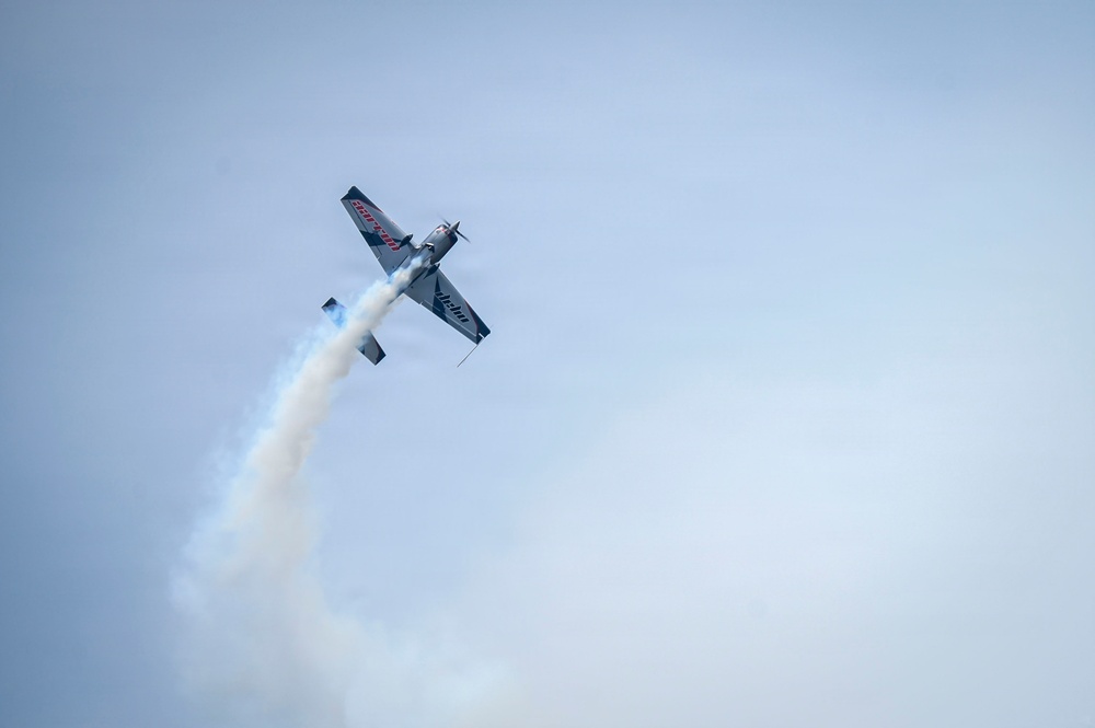 A single-engine aerobatic aircraft is performing a maneuver at an airshow, captured from a low angle looking upward. The aircraft appears to be inverted or in a steep bank, trailing a white smoke stream behind it. The overcast sky provides a pale blue-grey backdrop to the performance.
