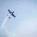 A single-engine aerobatic aircraft is performing a maneuver at an airshow, captured from a low angle looking upward. The aircraft appears to be inverted or in a steep bank, trailing a white smoke stream behind it. The overcast sky provides a pale blue-grey backdrop to the performance.