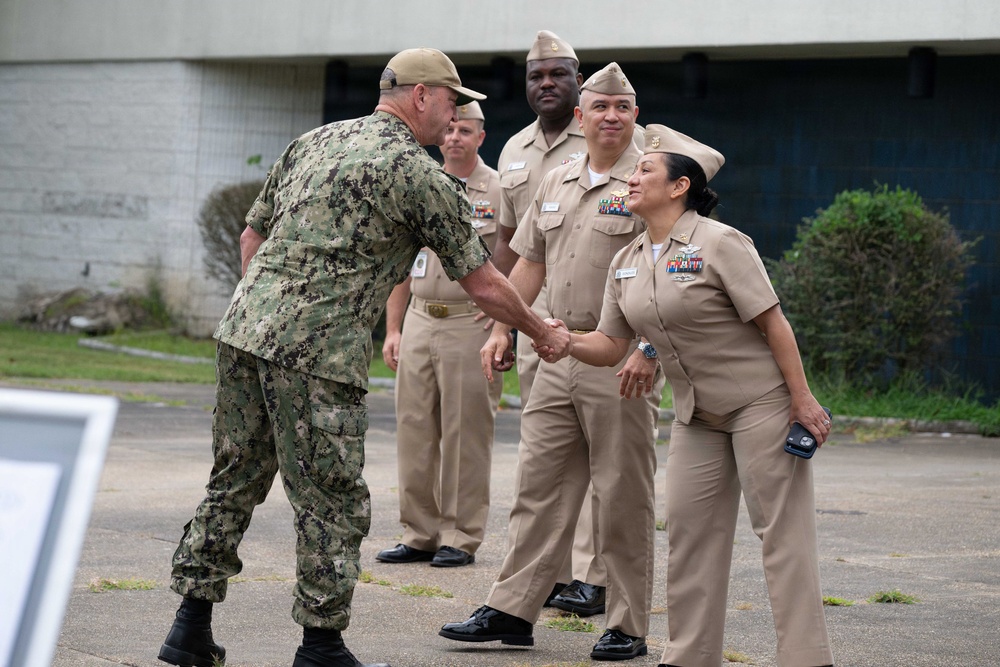 A U.S. Navy officer in digital camouflage (NWU Type III) is shaking hands with a female Navy officer in khaki service uniform (NWU Type II/Service Khaki). Several other Navy personnel in khaki uniforms are standing in formation or at ease in the background. The setting appears to be an outdoor military installation or base.
