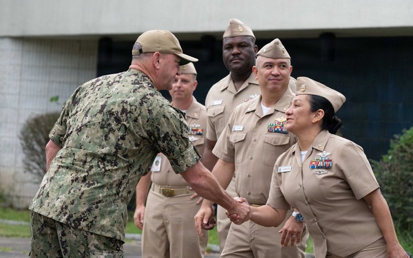 A U.S. Navy officer in digital camouflage (NWU Type III) is shaking hands with a female Navy officer in khaki service uniform (NWU Type II/Service Khaki). Several other Navy personnel in khaki uniforms are standing in formation or at ease in the background. The setting appears to be an outdoor military installation or base.