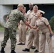 A U.S. Navy officer in digital camouflage (NWU Type III) is shaking hands with a female Navy officer in khaki service uniform (NWU Type II/Service Khaki). Several other Navy personnel in khaki uniforms are standing in formation or at ease in the background. The setting appears to be an outdoor military installation or base.