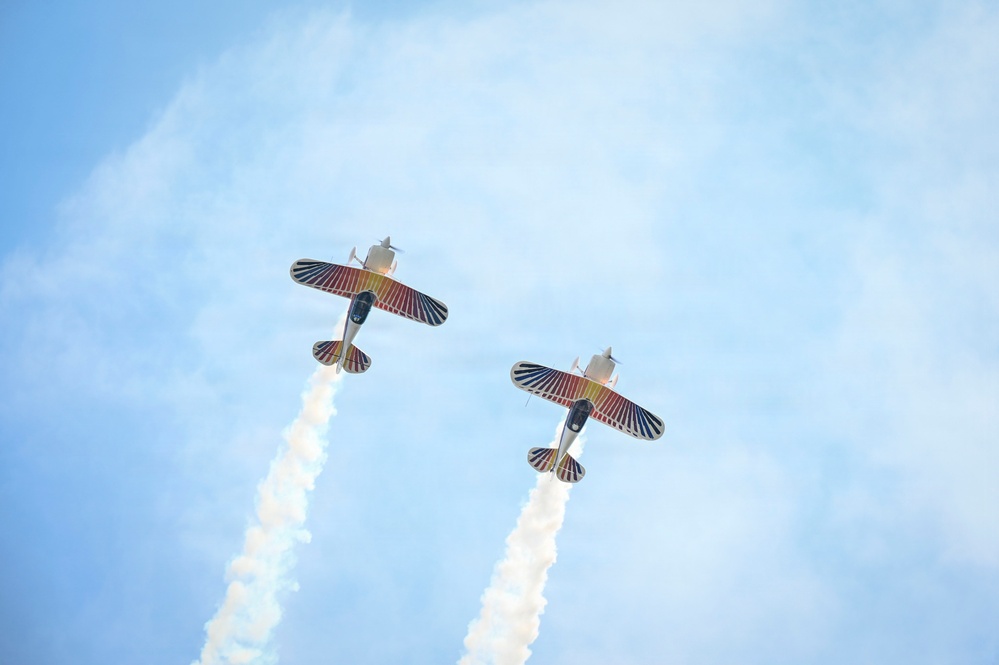 Two aerobatic biplanes are captured from below during an airshow performance, flying in close formation against a blue sky. Both aircraft display matching colorful livery with red, blue, and gold wing patterns and are trailing white smoke streams. The low-angle perspective emphasizes the underside of the aircraft and the parallel smoke trails.