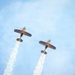 Two aerobatic biplanes are captured from below during an airshow performance, flying in close formation against a blue sky. Both aircraft display matching colorful livery with red, blue, and gold wing patterns and are trailing white smoke streams. The low-angle perspective emphasizes the underside of the aircraft and the parallel smoke trails.