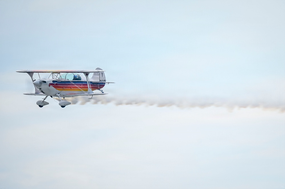 A white biplane with colorful horizontal stripes (red, orange, yellow, blue) is captured in flight against a pale blue-grey sky. The aircraft is emitting a white smoke trail extending horizontally behind it, suggesting an aerobatic or airshow performance. The registration number visible on the fuselage appears to read 'N219JB' or similar.