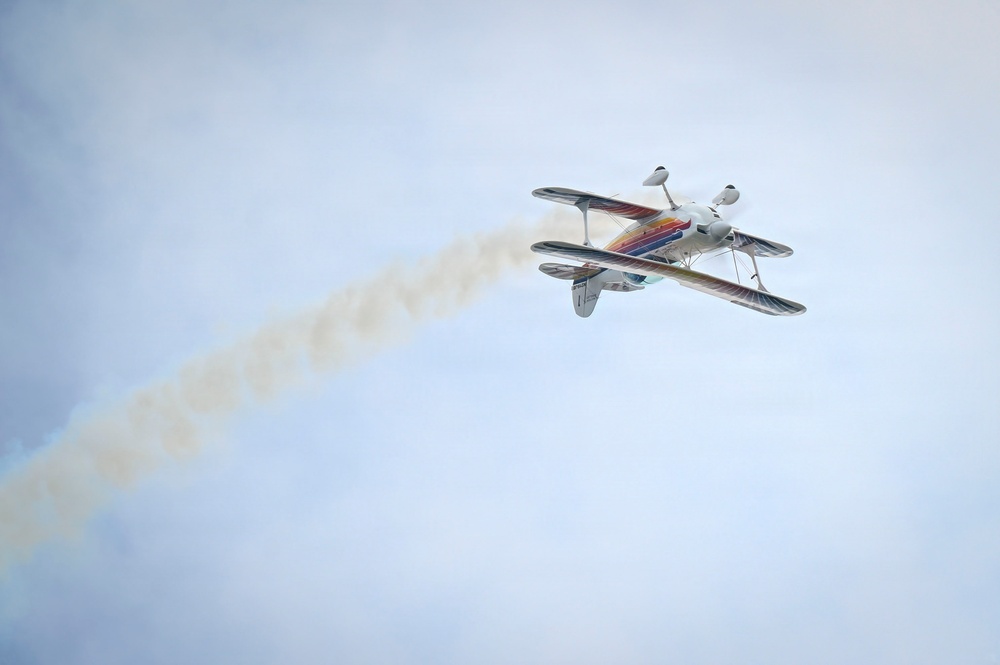 A colorful aerobatic biplane is captured mid-flight against an overcast sky, trailing a white smoke stream behind it. The aircraft appears to be performing an aerobatic maneuver, likely inverted or in a steep bank, during what appears to be an airshow. The smoke trail extends diagonally from the lower left to the upper right of the frame, emphasizing the aircraft's flight path.