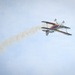 A colorful aerobatic biplane is captured mid-flight against an overcast sky, trailing a white smoke stream behind it. The aircraft appears to be performing an aerobatic maneuver, likely inverted or in a steep bank, during what appears to be an airshow. The smoke trail extends diagonally from the lower left to the upper right of the frame, emphasizing the aircraft's flight path.