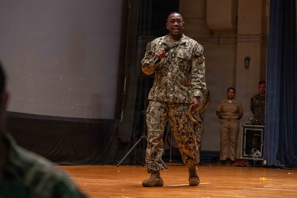A U.S. Navy servicemember in NWU Type III (woodland digital camouflage) uniform is speaking into a microphone on a wooden stage, appearing to address an audience. The speaker is smiling and holding a cover (hat) in one hand while gesturing with the microphone hand. Two additional military personnel are visible standing in the background near the stage.