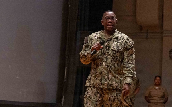 A U.S. Navy servicemember in NWU Type III (woodland digital camouflage) uniform is speaking into a microphone on a wooden stage, appearing to address an audience. The speaker is smiling and holding a cover (hat) in one hand while gesturing with the microphone hand. Two additional military personnel are visible standing in the background near the stage.