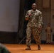 A U.S. Navy servicemember in NWU Type III (woodland digital camouflage) uniform is speaking into a microphone on a wooden stage, appearing to address an audience. The speaker is smiling and holding a cover (hat) in one hand while gesturing with the microphone hand. Two additional military personnel are visible standing in the background near the stage.