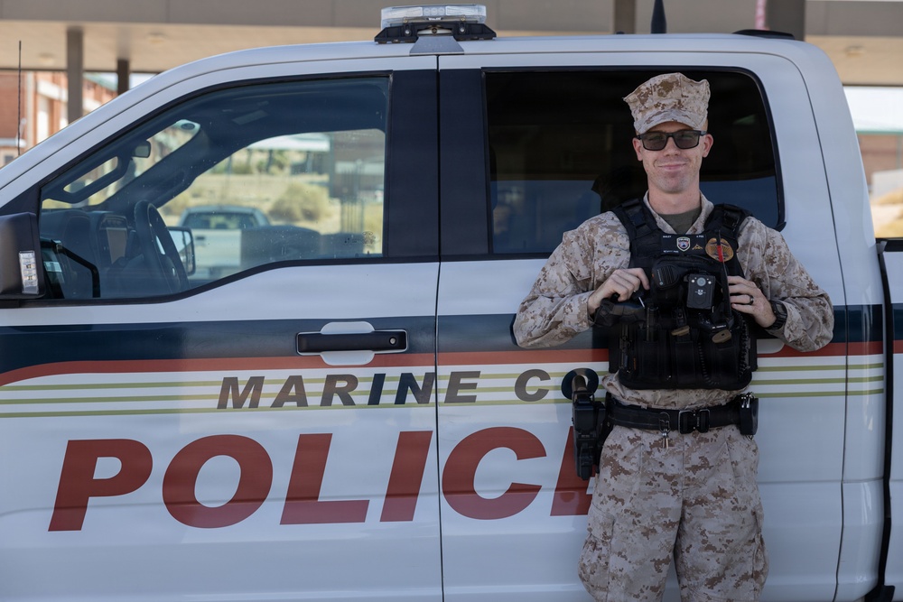 A uniformed Marine Corps Police officer stands in front of a white Marine Corps Police vehicle. The officer is wearing desert camouflage (MARPAT) uniform with a tactical vest and is holding what appears to be a body camera or radio equipment. The vehicle door clearly displays 'MARINE CO[RPS] POLICE' in large red lettering.