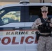 A uniformed Marine Corps Police officer stands in front of a white Marine Corps Police vehicle. The officer is wearing desert camouflage (MARPAT) uniform with a tactical vest and is holding what appears to be a body camera or radio equipment. The vehicle door clearly displays 'MARINE CO[RPS] POLICE' in large red lettering.