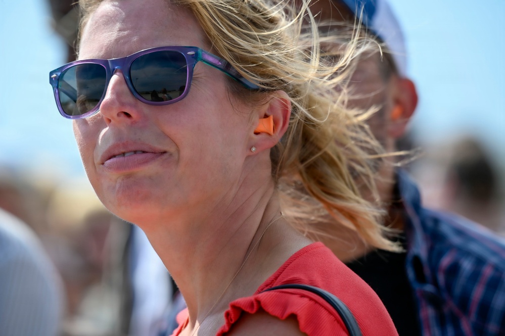 A close-up photograph of a blonde woman wearing purple sunglasses and an orange foam earplug in her visible ear, suggesting she is at a loud outdoor event. Her hair is blowing in the wind against a bright blue sky background. She appears to be watching something in the distance, with other people visible but blurred behind her.