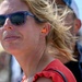 A close-up photograph of a blonde woman wearing purple sunglasses and an orange foam earplug in her visible ear, suggesting she is at a loud outdoor event. Her hair is blowing in the wind against a bright blue sky background. She appears to be watching something in the distance, with other people visible but blurred behind her.