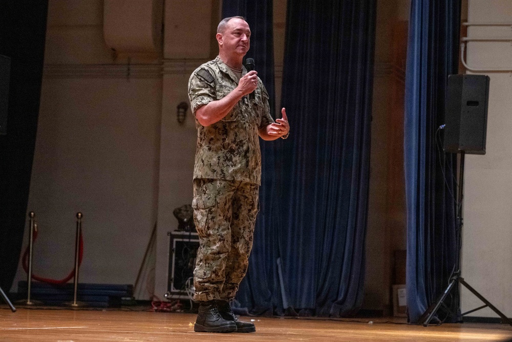 A military personnel in digital camouflage uniform is standing on a stage speaking into a handheld microphone. The setting appears to be an auditorium or theater with blue curtains in the background and a speaker system visible. The individual appears to be addressing an audience during a formal military presentation or briefing.