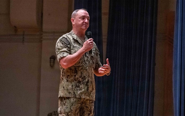 A military personnel in digital camouflage uniform is standing on a stage speaking into a handheld microphone. The setting appears to be an auditorium or theater with blue curtains in the background and a speaker system visible. The individual appears to be addressing an audience during a formal military presentation or briefing.