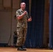 A military personnel in digital camouflage uniform is standing on a stage speaking into a handheld microphone. The setting appears to be an auditorium or theater with blue curtains in the background and a speaker system visible. The individual appears to be addressing an audience during a formal military presentation or briefing.