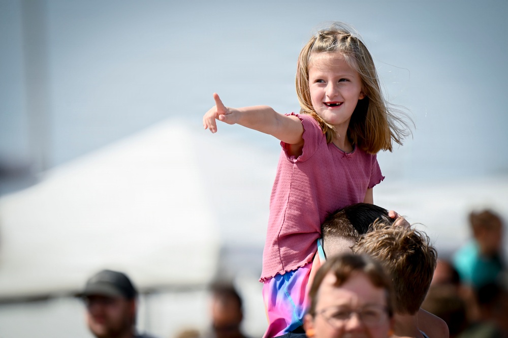 A young girl with shoulder-length blonde hair sits on an adult's shoulders at what appears to be an outdoor public event or festival. She is smiling and pointing enthusiastically at something off-camera, wearing a pink top and colorful shorts. A white event tent and a blurred crowd are visible in the background.
