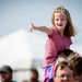 A young girl with shoulder-length blonde hair sits on an adult's shoulders at what appears to be an outdoor public event or festival. She is smiling and pointing enthusiastically at something off-camera, wearing a pink top and colorful shorts. A white event tent and a blurred crowd are visible in the background.