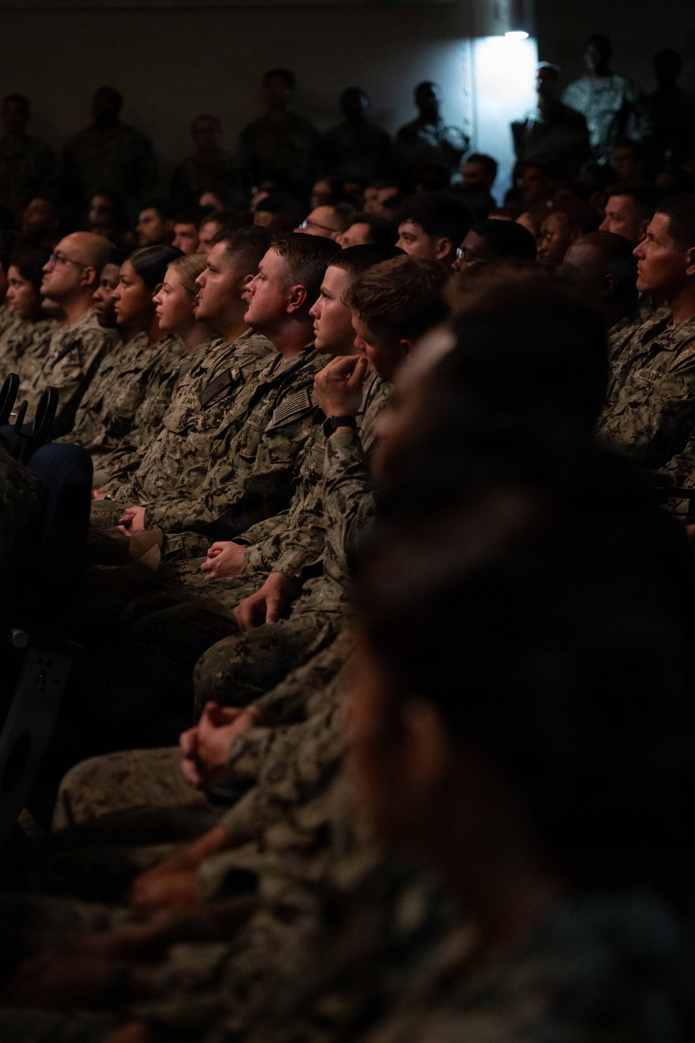 A large group of military personnel in camouflage uniforms are seated in rows, attentively facing a common direction, suggesting they are attending a briefing, presentation, or ceremony. The audience includes both male and female service members wearing what appears to be U.S. Navy NWU (Naval Working Uniform) and possibly Army ACU patterns. The venue is dimly lit with a single bright light source visible in the background.