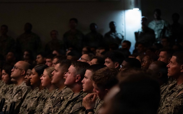 A large group of military personnel in camouflage uniforms are seated in rows, attentively facing a common direction, suggesting they are attending a briefing, presentation, or ceremony. The audience includes both male and female service members wearing what appears to be U.S. Navy NWU (Naval Working Uniform) and possibly Army ACU patterns. The venue is dimly lit with a single bright light source visible in the background.