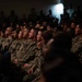 A large group of military personnel in camouflage uniforms are seated in rows, attentively facing a common direction, suggesting they are attending a briefing, presentation, or ceremony. The audience includes both male and female service members wearing what appears to be U.S. Navy NWU (Naval Working Uniform) and possibly Army ACU patterns. The venue is dimly lit with a single bright light source visible in the background.